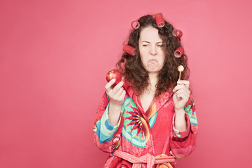 .Close up portrait of grimacing housewife in colorful bathrobe feeling stressed to eat healthy organic apple or unhealthy candy, dislike to keep diet, isolated on pink studio wall with copy space.