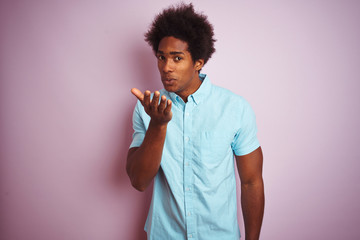 Young american man with afro hair wearing blue shirt standing over isolated pink background looking at the camera blowing a kiss with hand on air being lovely and sexy. Love expression.