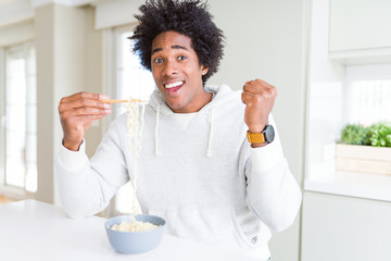 African American man eating asian noodles using chopsticks at home screaming proud and celebrating victory and success very excited, cheering emotion