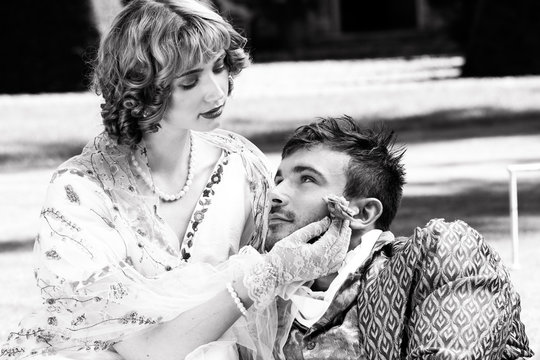 Young lovers dressed in vintage clothing. Man looking into partner's eyes as she strokes his face with a flower