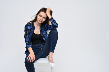 Portrait of a cute brunette girl, a young woman with beautiful curly hair in a blue shirt and jeans, is sitting on a chair on a white background. Smiling, talking with emotions, showing hands.