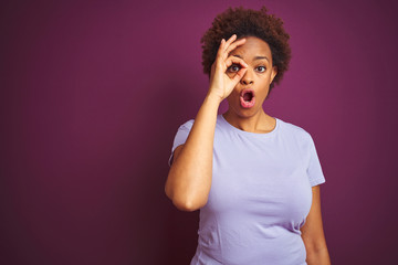 Young beautiful african american woman with afro hair over isolated purple background doing ok gesture shocked with surprised face, eye looking through fingers. Unbelieving expression.