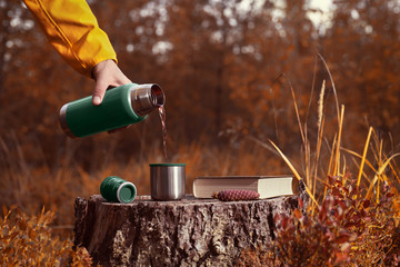 A man pours hot tea from a thermos into a mug standing on a stump in the autumn forest. Nearby lies...