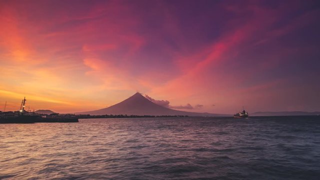 Magayon volcano of perfect symmetric conical shape by sea with boats under orange morning sky time lapse. Concept travel paradise tourism. Exotic landscape. Bicol Region Luzon island Philippines. 4K