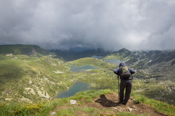 Hiker with blue jacket with hood over head, walking sticks and backpack standing at the edge of Lake peak and looking at seven Rila lakes through thick fog