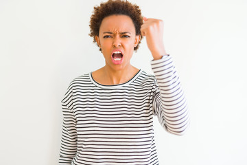 Young beautiful african american woman wearing stripes sweater over white background angry and mad raising fist frustrated and furious while shouting with anger. Rage and aggressive concept.