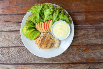 Top view grilled chicken breast steak with sliced avocado, red apple and green lettuce leaves served with salad dressing in white bowl on dish in cafe and restaurant.