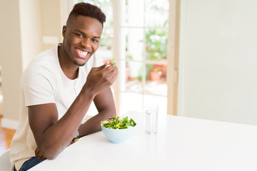 Fototapeta na wymiar Handsome african young man eating a healthy vegetable salad using a fork to eat lettuce, happy and smiling sitting on the table