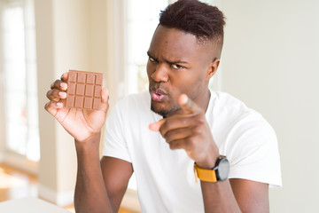 Young african american man eating chocolate bar pointing with finger to the camera and to you, hand...