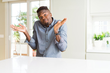 Handsome african american man on white table clueless and confused expression with arms and hands...