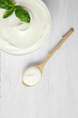 Bowl of fresh sour cream with basil and spoon on white wooden table, flat lay