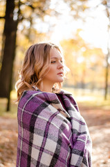 Young beautiful blonde woman wrapped in checkered blanket or plaid walks outdoors through the autumn park at sunset. Comfort and cosiness concept.