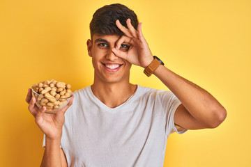 Young indian man holding bowl with peanuts over isolated yellow background with happy face smiling doing ok sign with hand on eye looking through fingers