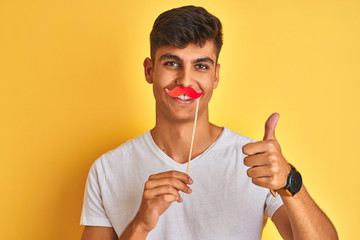 Young indian man holding fanny mustache standing over isolated yellow background happy with big...