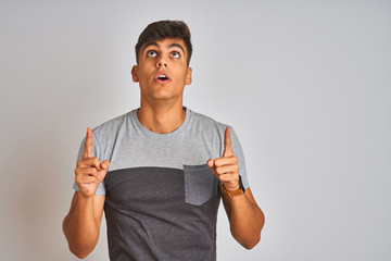 Young indian man wearing casual t-shirt standing over isolated white background amazed and surprised looking up and pointing with fingers and raised arms.