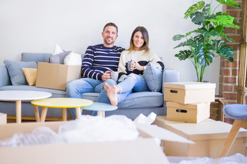 Fototapeta na wymiar Young beautiful couple sitting on the sofa drinking cup of coffee at new home around cardboard boxes