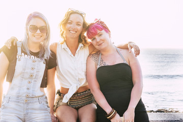 group of three women hugged and sitted at the beach looking and smiling at the camera with the sea and the blue water at the background - friendship goals concept