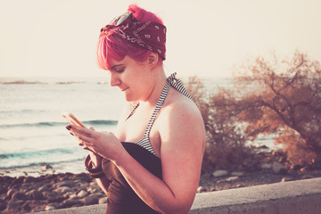 woman at the beach at the sunset with the blue sea and white sky at the background, typing and chatting with her phone - online lifestyle concept