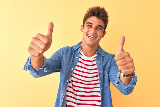Young handsome man wearing striped t-shirt and denim shirt over isolated yellow background approving doing positive gesture with hand, thumbs up smiling and happy for success. Winner gesture.