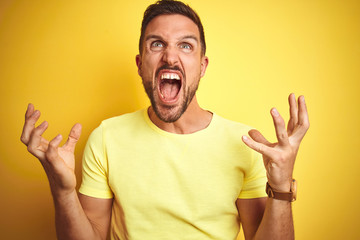 Young handsome man wearing casual yellow t-shirt over yellow isolated background crazy and mad shouting and yelling with aggressive expression and arms raised. Frustration concept.