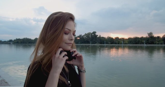 Young woman puts on large wireless headphones and listens to favourite music on streaming platform at sunset