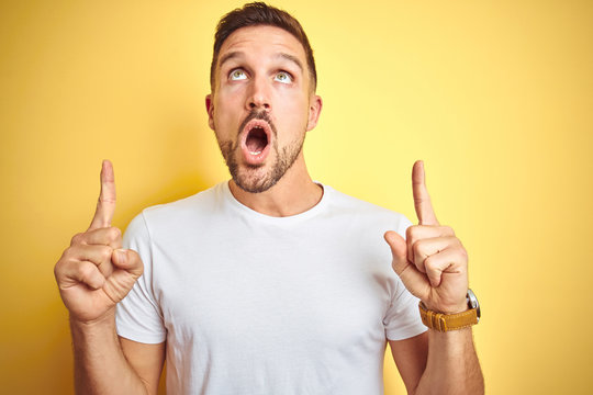 Young handsome man wearing casual white t-shirt over yellow isolated background amazed and surprised looking up and pointing with fingers and raised arms.