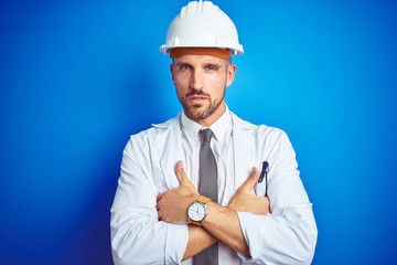 Young handsome engineer man wearing safety helmet over blue isolated background skeptic and nervous, disapproving expression on face with crossed arms. Negative person.