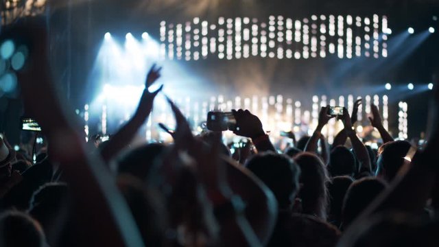 A numerous audience at a music performance, shooting it on smartphones and clapping hands, we see back silhouettes