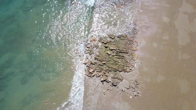 Scenic vertical aerial view of flight along remote coast, with calm turquoise ocean waves, sunlight reflection on wet sandy beach as background and copy space.