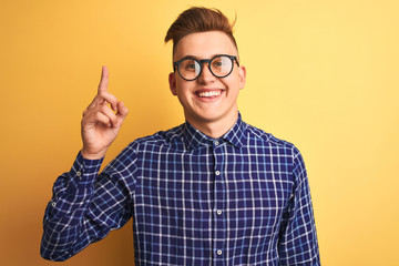 Fototapeta premium Young handsome man wearing casual shirt and glasses over isolated yellow background pointing finger up with successful idea. Exited and happy. Number one.