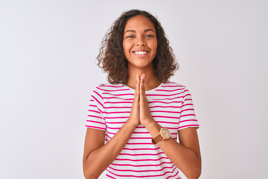 Young brazilian woman wearing pink striped t-shirt standing over isolated white background praying with hands together asking for forgiveness smiling confident.