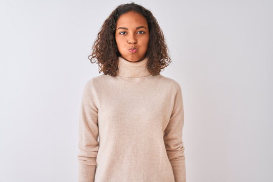 Young brazilian woman wearing turtleneck sweater standing over isolated white background puffing cheeks with funny face. Mouth inflated with air, crazy expression.