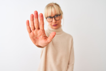 Middle age woman wearing turtleneck sweater and glasses over isolated white background doing stop sing with palm of the hand. Warning expression with negative and serious gesture on the face.