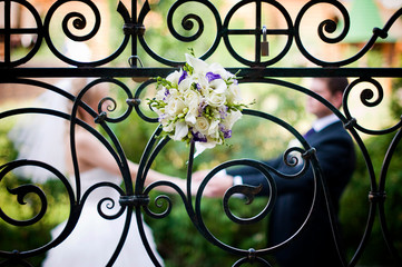 Fototapeta na wymiar A bride's bouquet is inserted into an elegant wrought-iron fence, behind which the newlyweds hold hands in defocus. grain