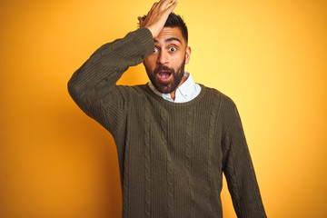 Young indian man wearing green sweater and shirt standing over isolated yellow background surprised with hand on head for mistake, remember error. Forgot, bad memory concept.