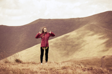 Girl hiker showing thumbs up while standing on the background of adorable mountains. Toned.