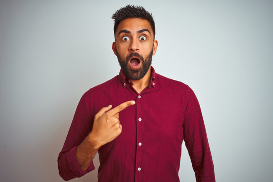 Young indian man wearing red elegant shirt standing over isolated grey background Surprised pointing with finger to the side, open mouth amazed expression.