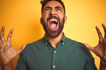 Young indian man wearing green shirt standing over isolated yellow background crazy and mad shouting and yelling with aggressive expression and arms raised. Frustration concept.