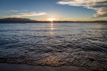 Smooth glassy water reflects with the golden sun setting behind flat mountains in the distance looking from Lakeside Beach at Lake Tahoe