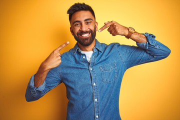 Young indian man wearing denim shirt standing over isolated yellow background smiling cheerful showing and pointing with fingers teeth and mouth. Dental health concept.