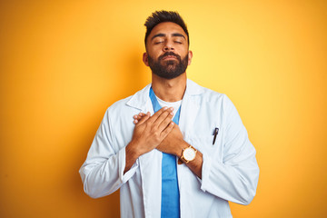 Young indian doctor man standing over isolated yellow background smiling with hands on chest with closed eyes and grateful gesture on face. Health concept.