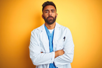 Young indian doctor man standing over isolated yellow background skeptic and nervous, disapproving expression on face with crossed arms. Negative person.