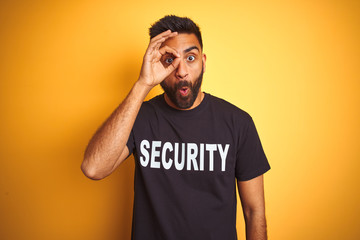 Arab indian hispanic safeguard man wearing security uniform over isolated yellow background doing ok gesture shocked with surprised face, eye looking through fingers. Unbelieving expression.