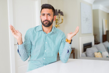 Handsome hispanic man at home clueless and confused expression with arms and hands raised. Doubt...