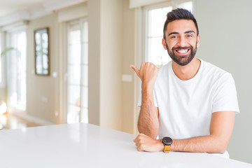 Handsome hispanic man casual white t-shirt at home smiling with happy face looking and pointing to the side with thumb up.