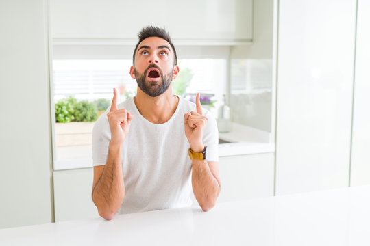 Handsome hispanic man casual white t-shirt at home amazed and surprised looking up and pointing with fingers and raised arms.