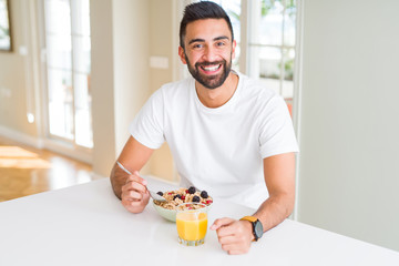 Handsome hispanic man eating healthy breakfast in the morning at home with a happy face standing and smiling with a confident smile showing teeth