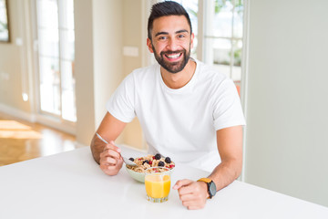 Handsome hispanic man eating healthy breakfast in the morning at home with a happy face standing...