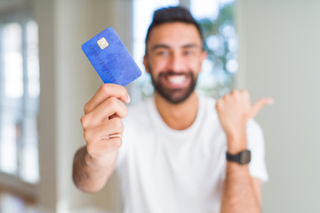 Handsome hispanic man holding credit card pointing and showing with thumb up to the side with happy face smiling