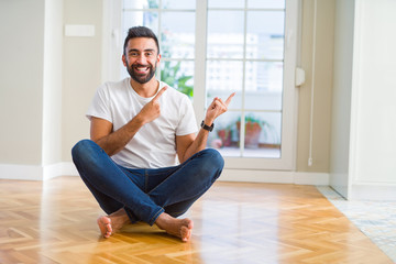 Handsome hispanic man wearing casual t-shirt sitting on the floor at home smiling and looking at the camera pointing with two hands and fingers to the side.